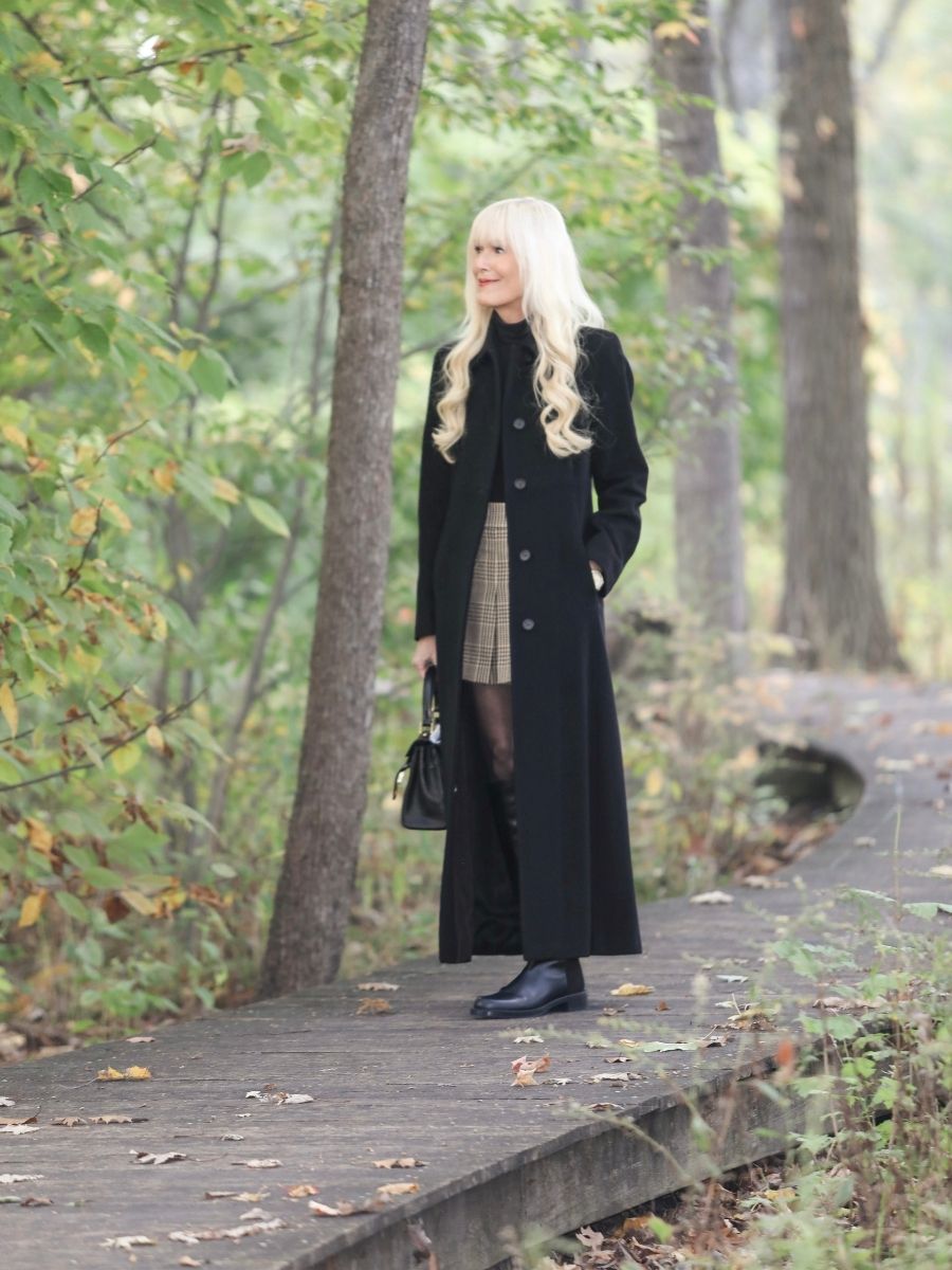 Woman wearing a long black coat with a plaid skirt and black ankle boots, standing on a wooded path in early fall.