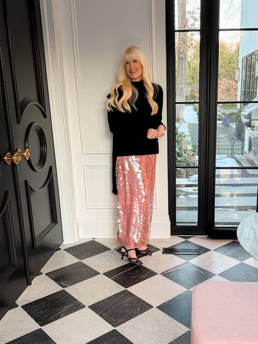 Woman wearing a black long-sleeve sweater with a pink sequin skirt and black heels, standing indoors by glass doors.