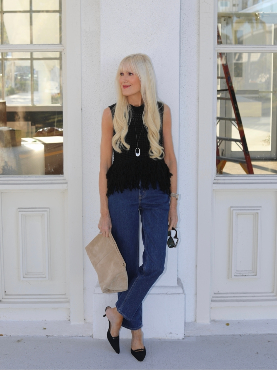 Blonde woman wearing a sleeveless black textured top with a fringe hem, straight-leg blue jeans, and black pointed-toe heels, holding a beige suede clutch while standing against a white building facade.