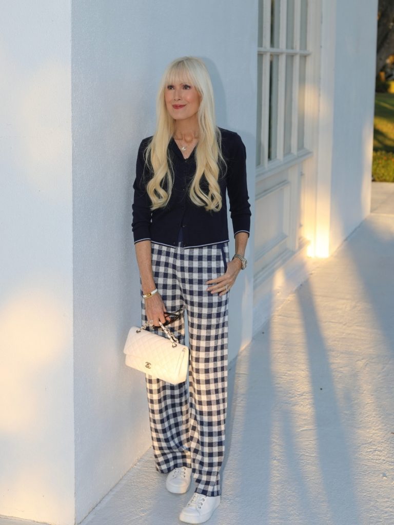 Woman wearing a navy knit cardigan layered over a button down with navy and white gingham wide leg pants, white leather sneakers, and a white quilted chain handbag, standing against a white building at golden hour.