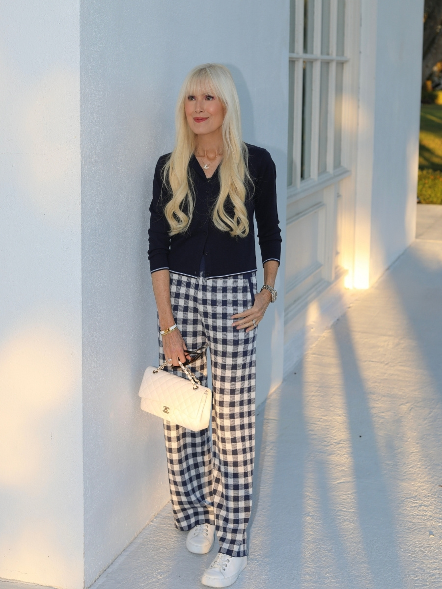 Woman wearing a navy knit cardigan layered over a button down with navy and white gingham wide leg pants, white leather sneakers, and a white quilted chain handbag, standing against a white building at golden hour.