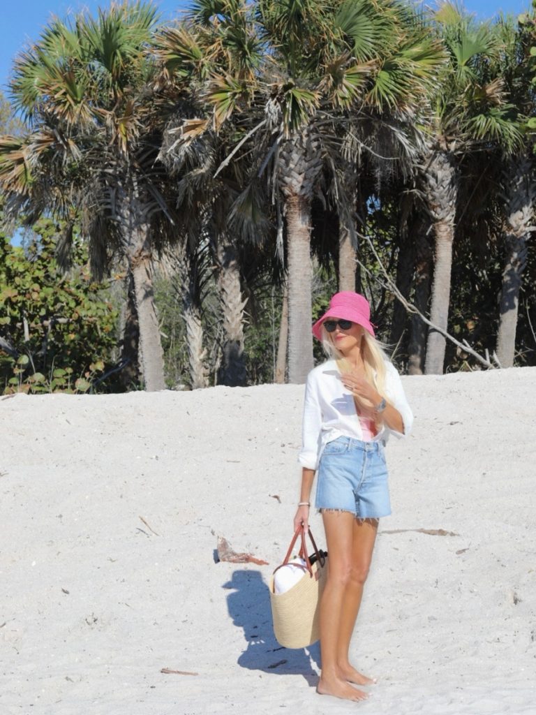 Woman in white button-down shirt and denim shorts standing barefoot on a sandy beach with palm trees, wearing a pink bucket hat and carrying a woven tote.