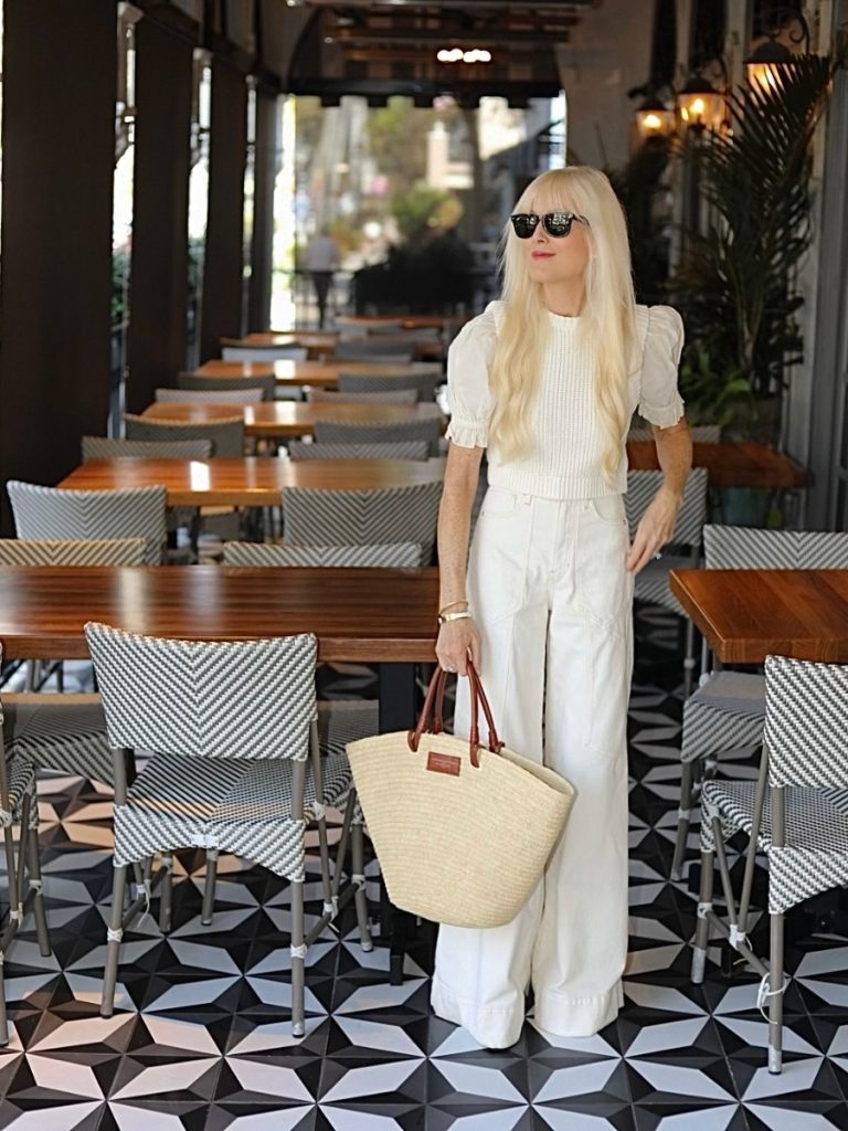 Mary Murnane standing at a restaurant in Naples wearing Veronica Beard Jeans, a Zara top, Ray-Ban Wayfarers and holding a Sézane Justine Basket Bag