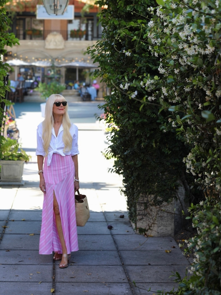 Woman wearing a white cotton button-down shirt tied at the waist and a pink and white striped linen midi skirt with a front slit, styled with neutral sandals and a raffia tote, walking through a sunlit garden courtyard.