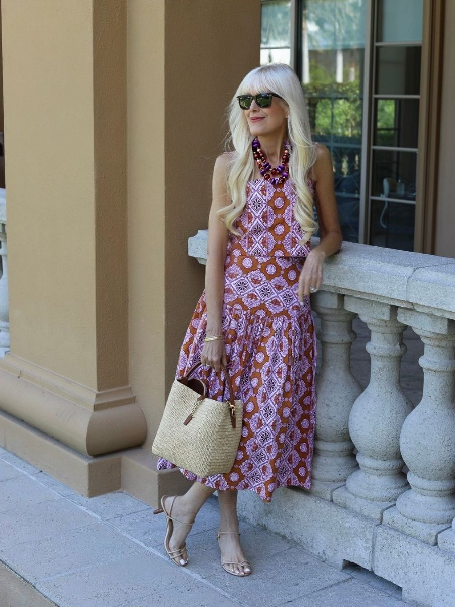 Woman in a patterned midi sundress with a vintage-inspired print, styled with neutral heeled sandals, a woven straw handbag, oversized beaded statement necklace, and black sunglasses, standing on a sunlit terrace.