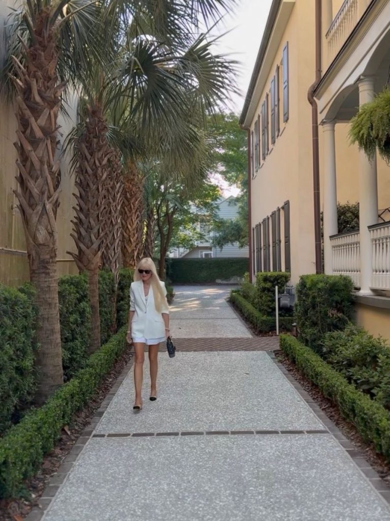Mary walking through a palm-lined Charleston pathway wearing a white Cinq à Sept blazer, Ralph Lauren linen shorts, Chanel pumps, and a Dior bag for an elegant summer date night outfit.