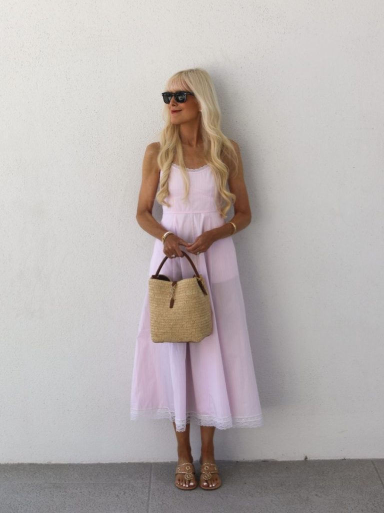 Mary Murnane wearing a soft blush pink midi dress with lace trim styled with a woven straw handbag, gold jewelry, black sunglasses, and minimalist sandals against a white wall. Feminine summer outfit inspiration with timeless modern classic style.