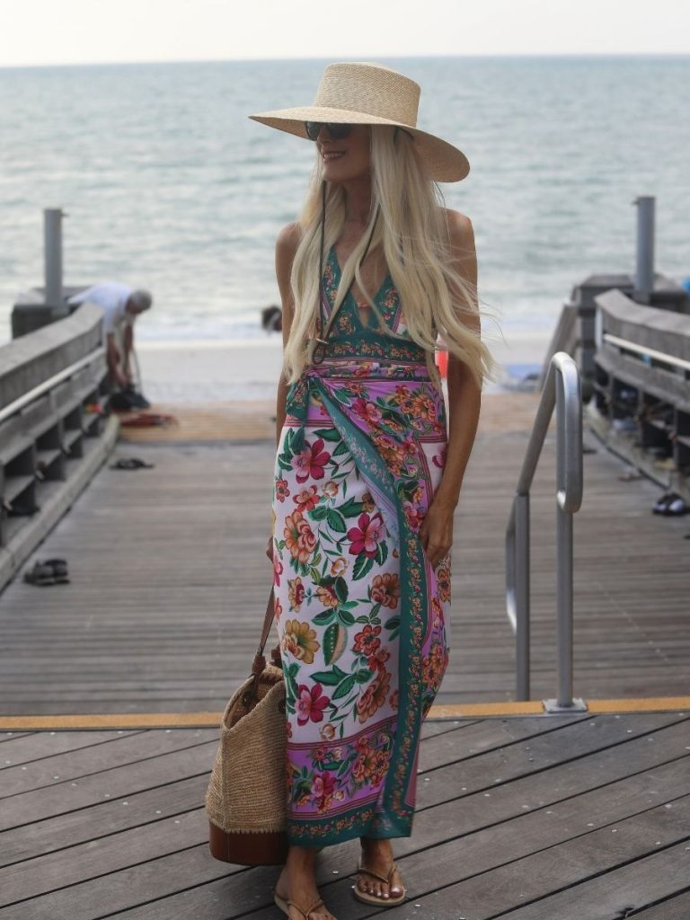 Woman wearing a floral swimsuit and matching pareo wrap skirt with a wide brim straw hat, woven beach bag, sandals, and sunglasses while standing on a beach boardwalk by the ocean.