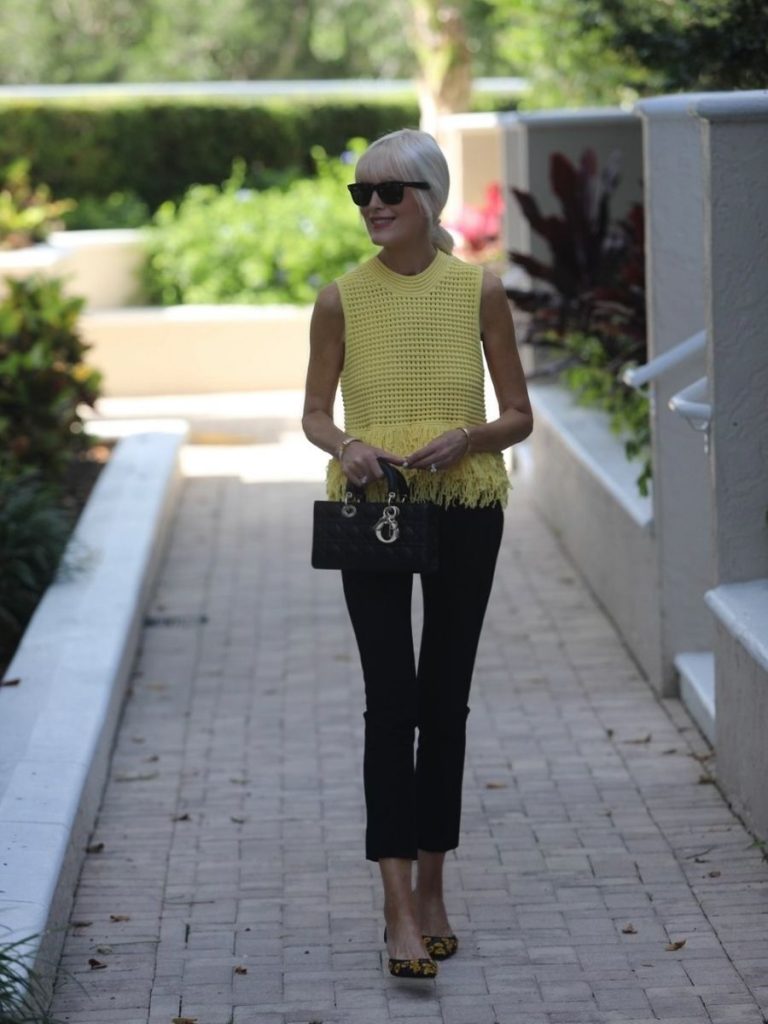 Mary Murnane wearing a yellow fringe knit sleeveless top with black tailored ankle pants and embroidered floral slingback heels in a tropical outdoor setting.