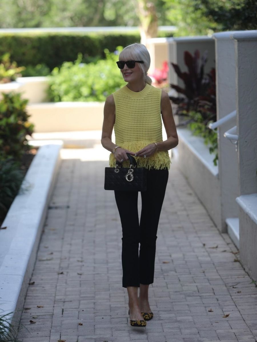 Mary Murnane wearing a yellow fringe knit sleeveless top with black tailored ankle pants and embroidered floral slingback heels in a tropical outdoor setting.