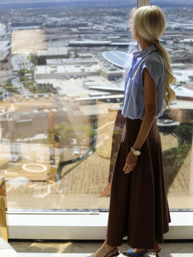 Woman wearing a light blue sleeveless button-front shirt tucked into a chocolate brown midi skirt while standing beside a large window overlooking the city.