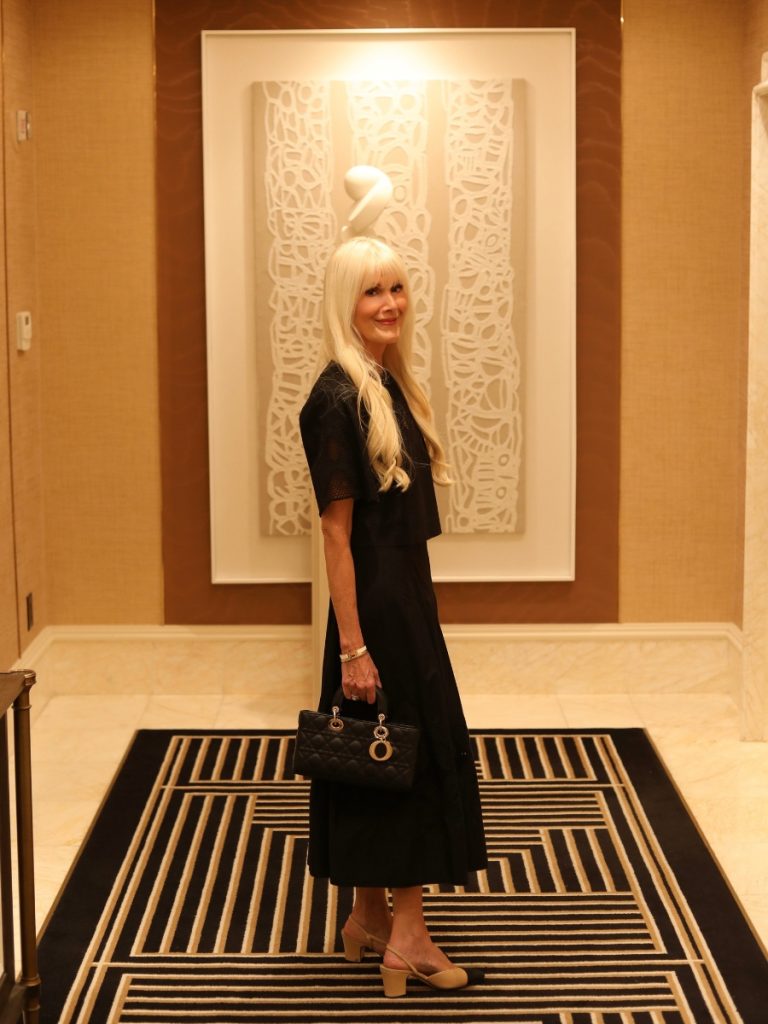 Woman wearing a black matching set with two-tone slingback heels and a structured black handbag in a sophisticated hotel interior.