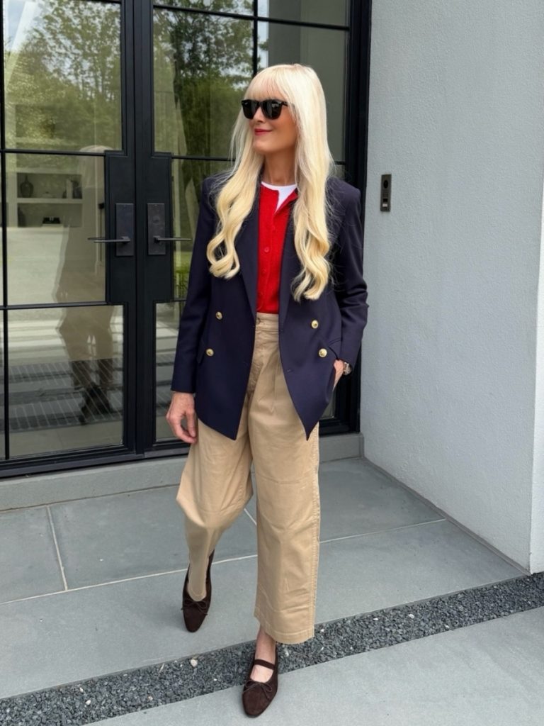 Woman wearing a navy double-breasted blazer layered over a bright red cardigan and white tee with cropped khaki trousers and brown flats, styled in a modern classic aesthetic outside a contemporary home.