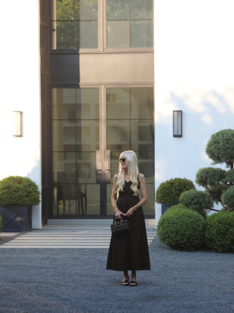 Mary Murnane wearing a black sleeveless maxi dress with a quilted Dior handbag, black strappy heels, and sunglasses, styled in front of a modern white home