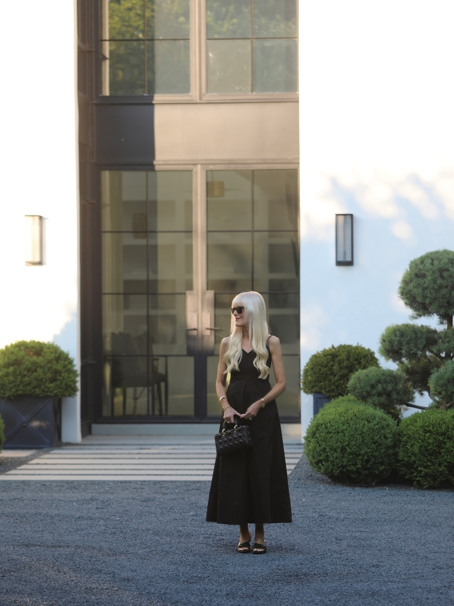 Mary Murnane wearing a black sleeveless maxi dress with a quilted Dior handbag, black strappy heels, and sunglasses, styled in front of a modern white home