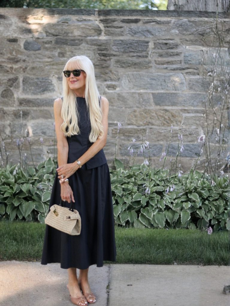 Mary wearing a timeless black summer outfit with a sleeveless tailored top, full midi skirt, neutral sandals, black sunglasses, and a woven top-handle bag.