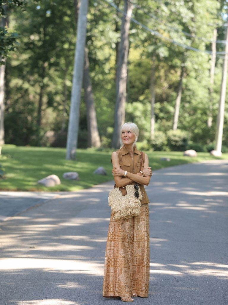 Blonde woman wearing a tan suede sleeveless zip-front top with a paisley-print wide-leg maxi skirt, standing on a tree-lined street holding a fringed straw fringe bag, modern classic summer outfit.