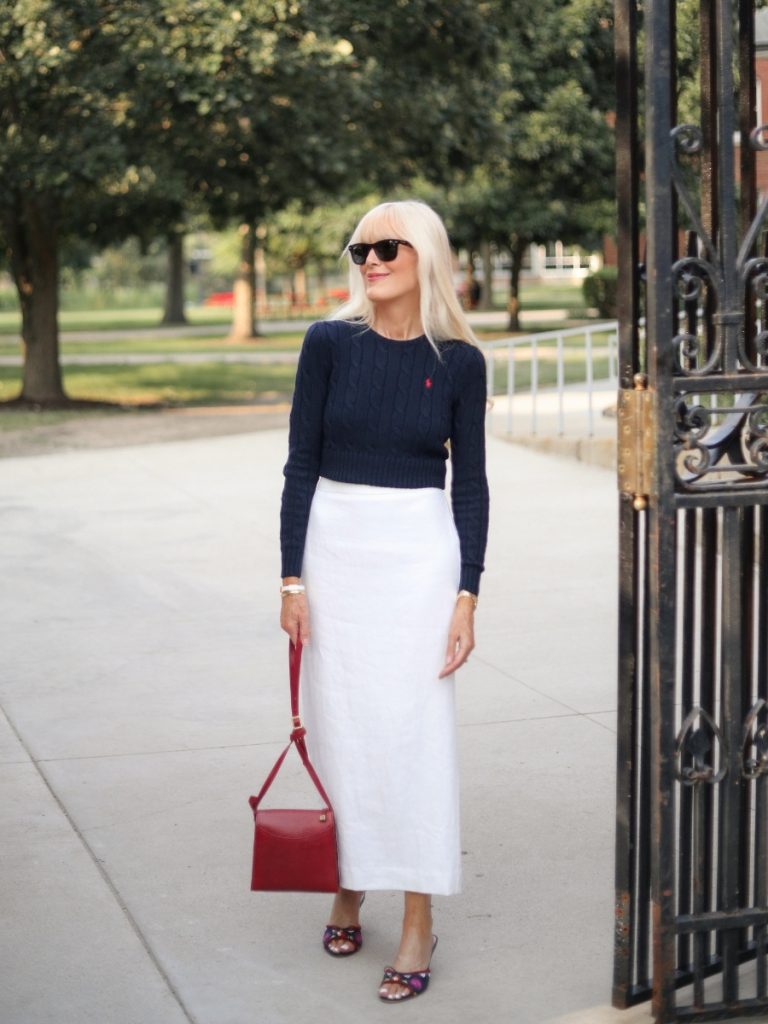 Mary wearing a white midi pencil skirt with a cropped navy cable-knit sweater, colorful sandals, black sunglasses, and a structured red handbag.