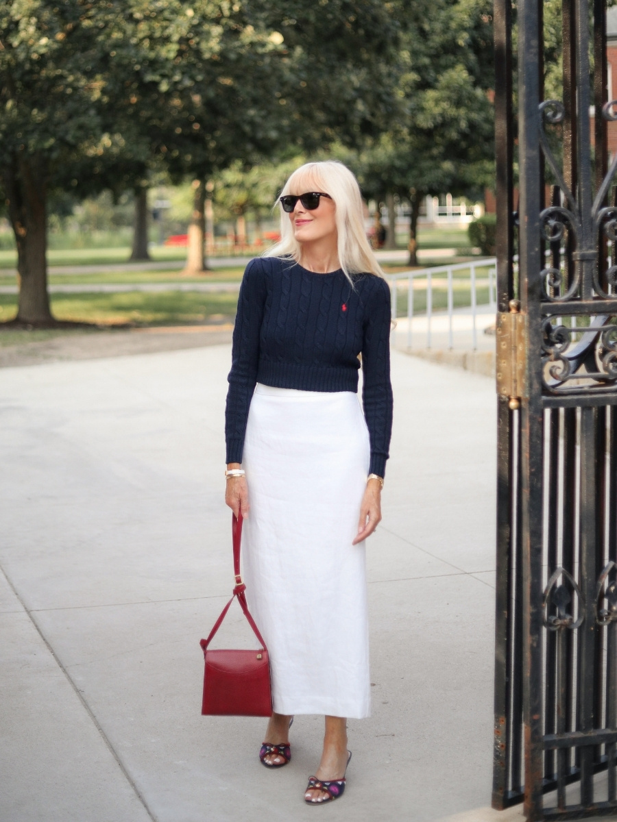 Mary wearing a white midi pencil skirt with a cropped navy cable-knit sweater, colorful sandals, black sunglasses, and a structured red handbag.