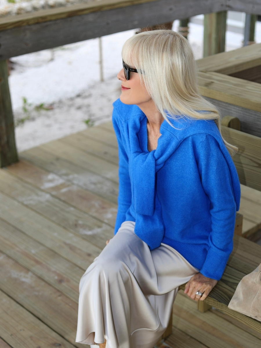 Woman wearing a cobalt blue sweater with a matching sweater draped over her shoulders, styled with an ivory satin midi skirt and black sunglasses.