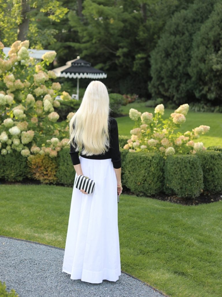 Mary Murnane wearing a black sweater over a white STAUD Wylie Maxi Dress styled as a skirt with a black and white striped clutch