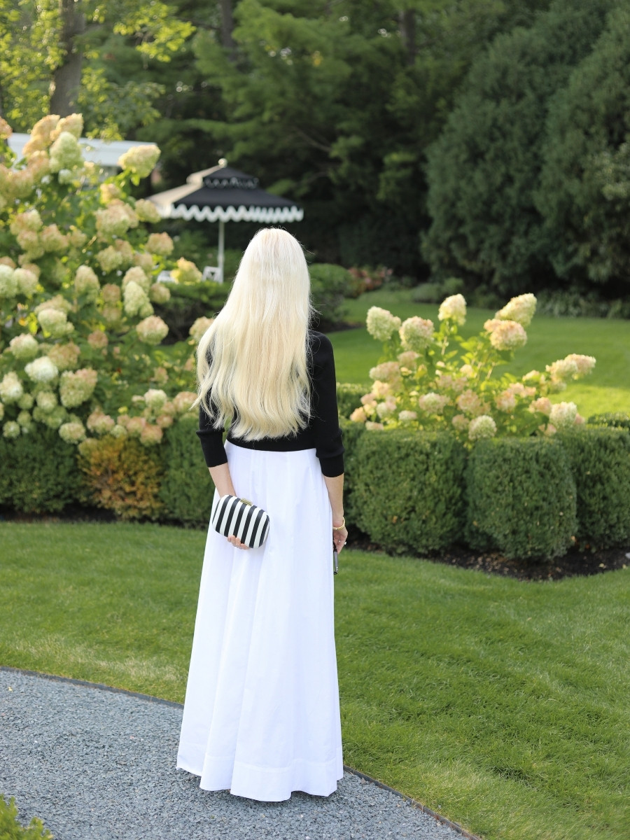 Mary Murnane wearing a black sweater over a white STAUD Wylie Maxi Dress styled as a skirt with a black and white striped clutch